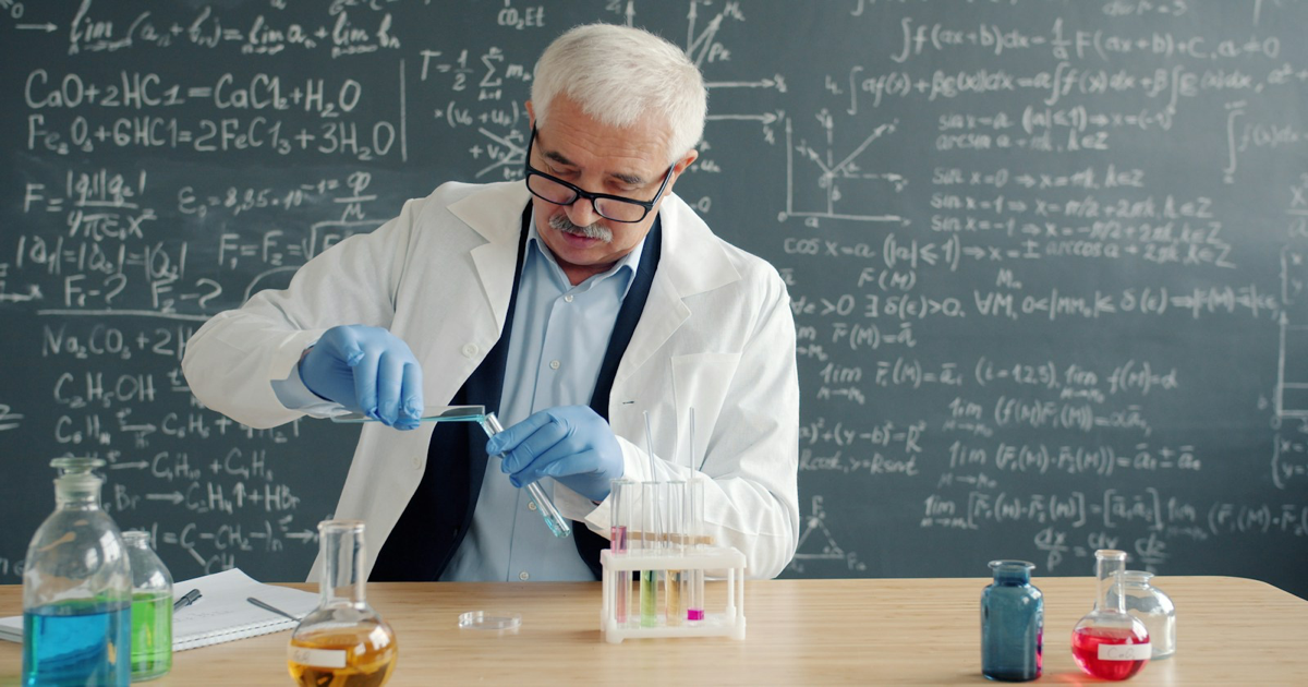 Photo: Vitaly Gariev / Unsplash (Unsplash License) A scientist in a white coat handling laboratory glassware beside a chalkboard.