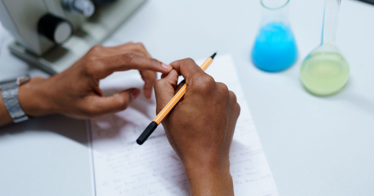 A person writes in a notebook beside laboratory glassware and a microscope.
