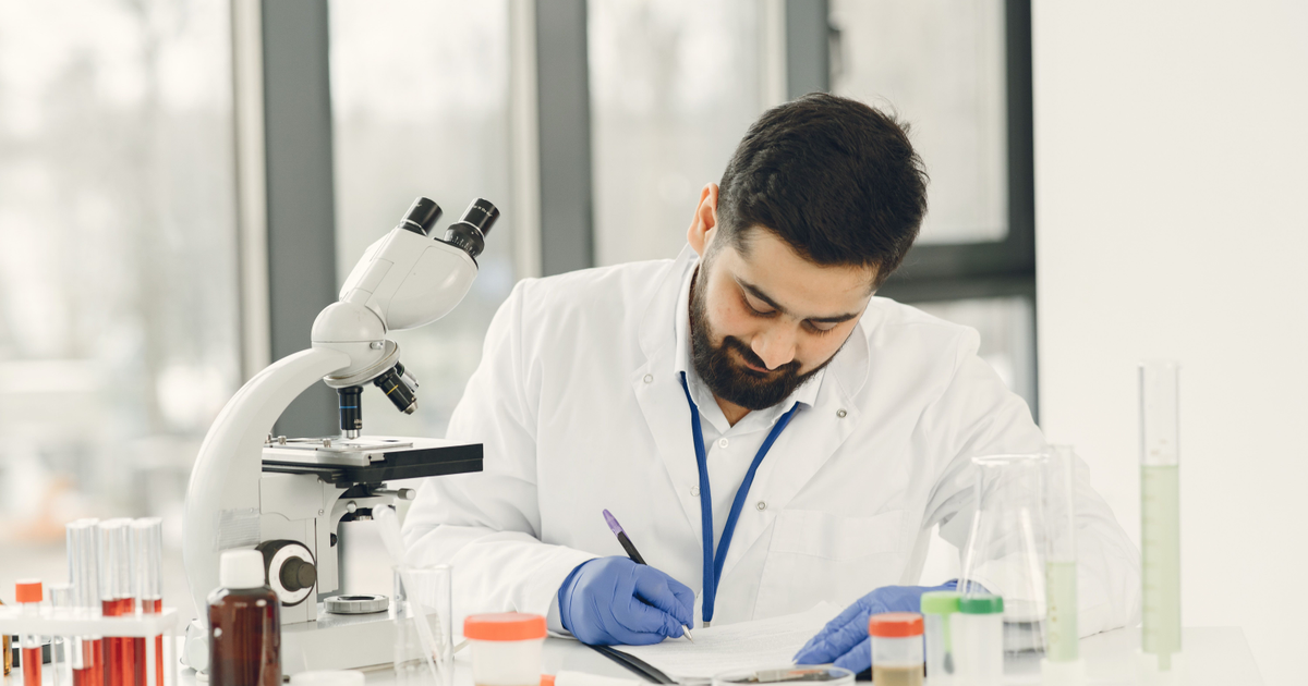A scientist writes on paper beside a microscope and laboratory glassware.