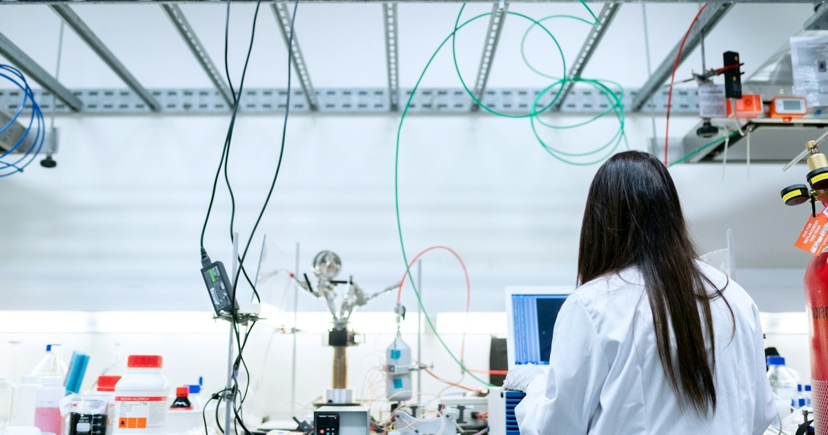 A scientist in a white coat works at a laptop in a laboratory.