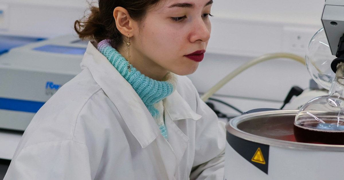 Photo: Faustina Okeke / Unsplash (Unsplash License) A scientist in a white laboratory coat working beside glass lab equipment.
