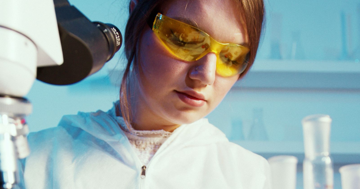 Photo: Artem Podrez / Pexels (Pexels License) A woman wearing yellow safety glasses beside a microscope and laboratory glassware.