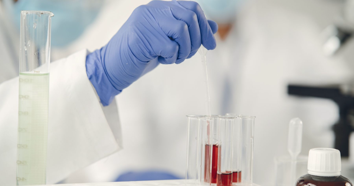 Photo: Gustavo Fring / Pexels (Pexels License) A gloved hand holds a pipette beside test tubes and laboratory glassware.
