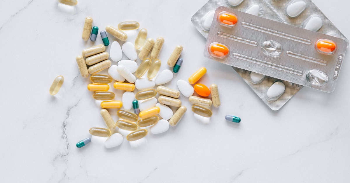 Loose capsules, fish-oil softgels and white tablets scattered on white marble beside two foil blister packs of pills